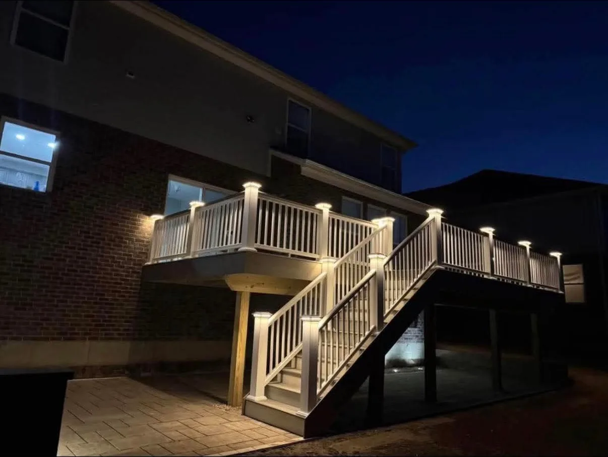 Elevated composite deck with white railing, lit post caps and stair lighting glowing over a paver patio at night