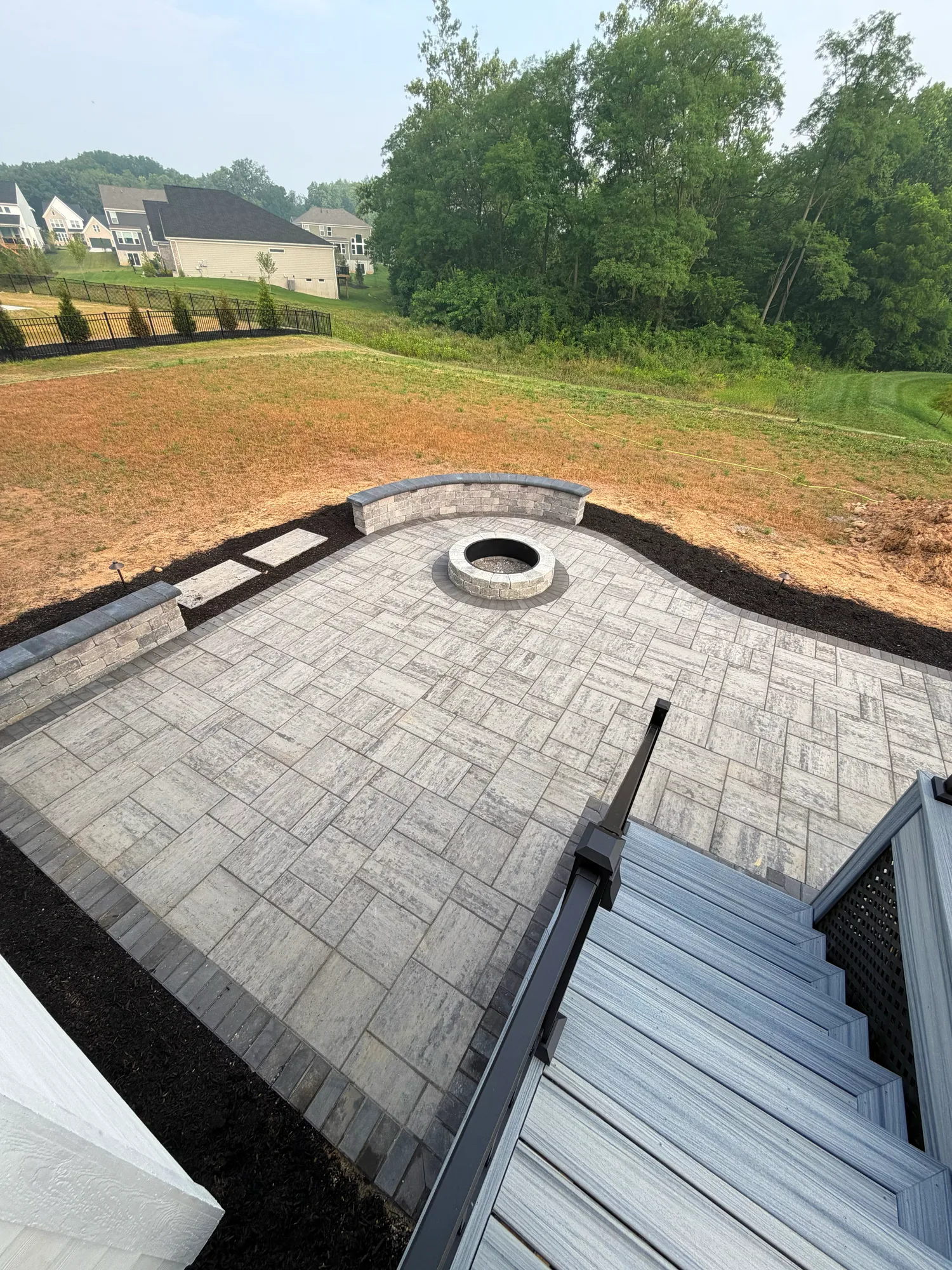 Paver patio with a round fire pit and composite deck steps at the edge of a graded yard