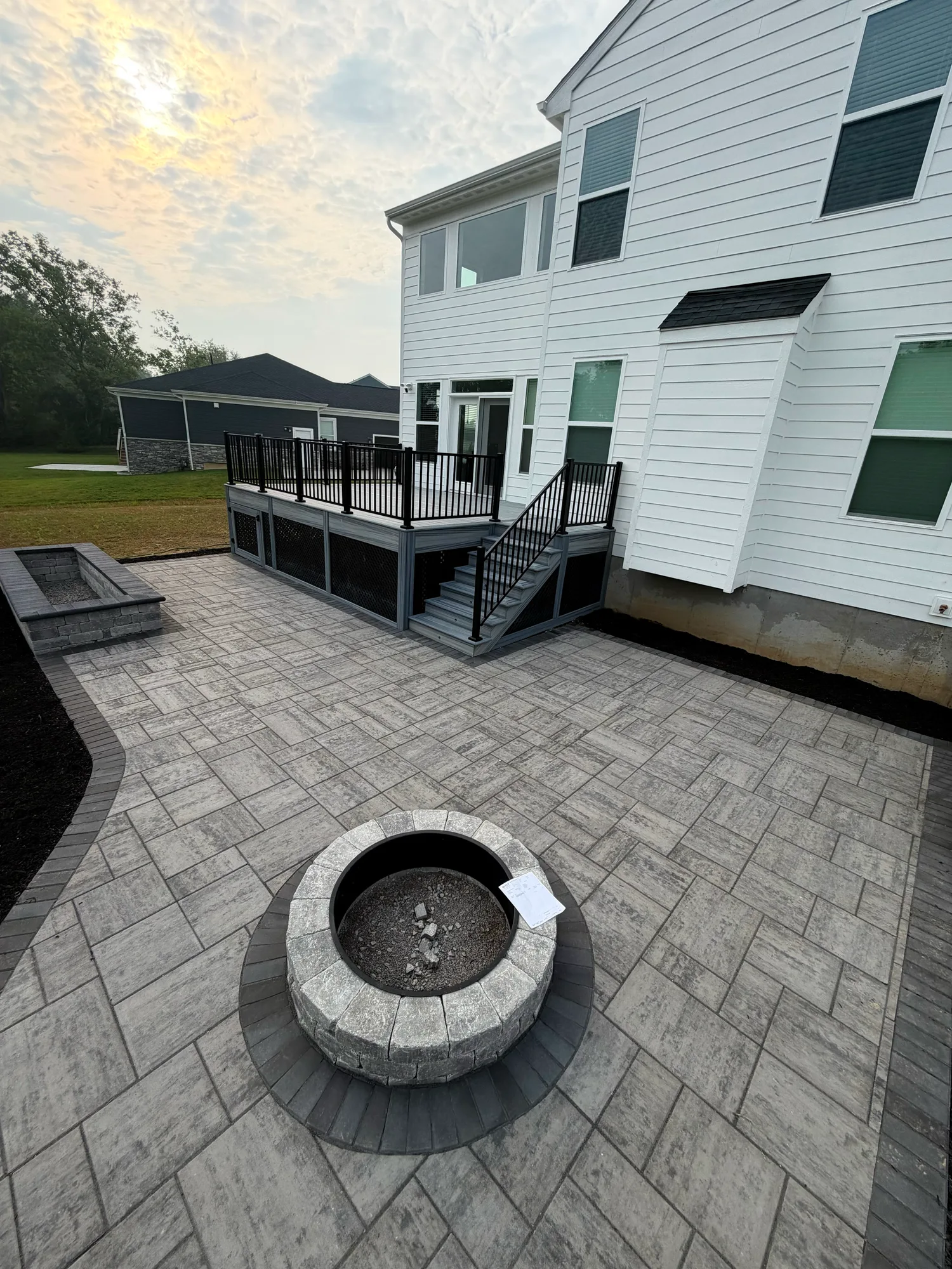 Stone fire pit on a large paver patio beside a black railed composite deck at a white sided home