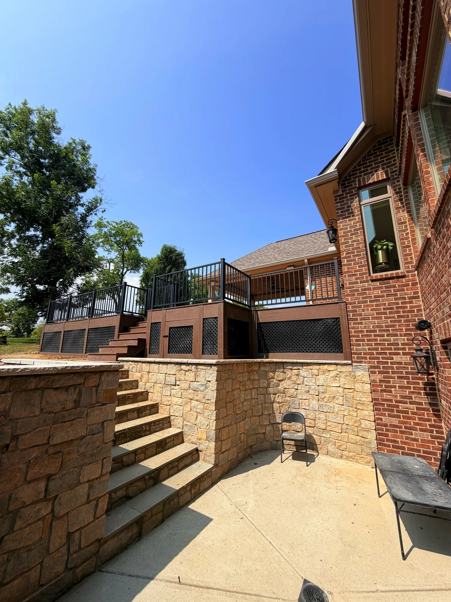 Raised composite deck with black railing above a stone patio and retaining wall at a brick home