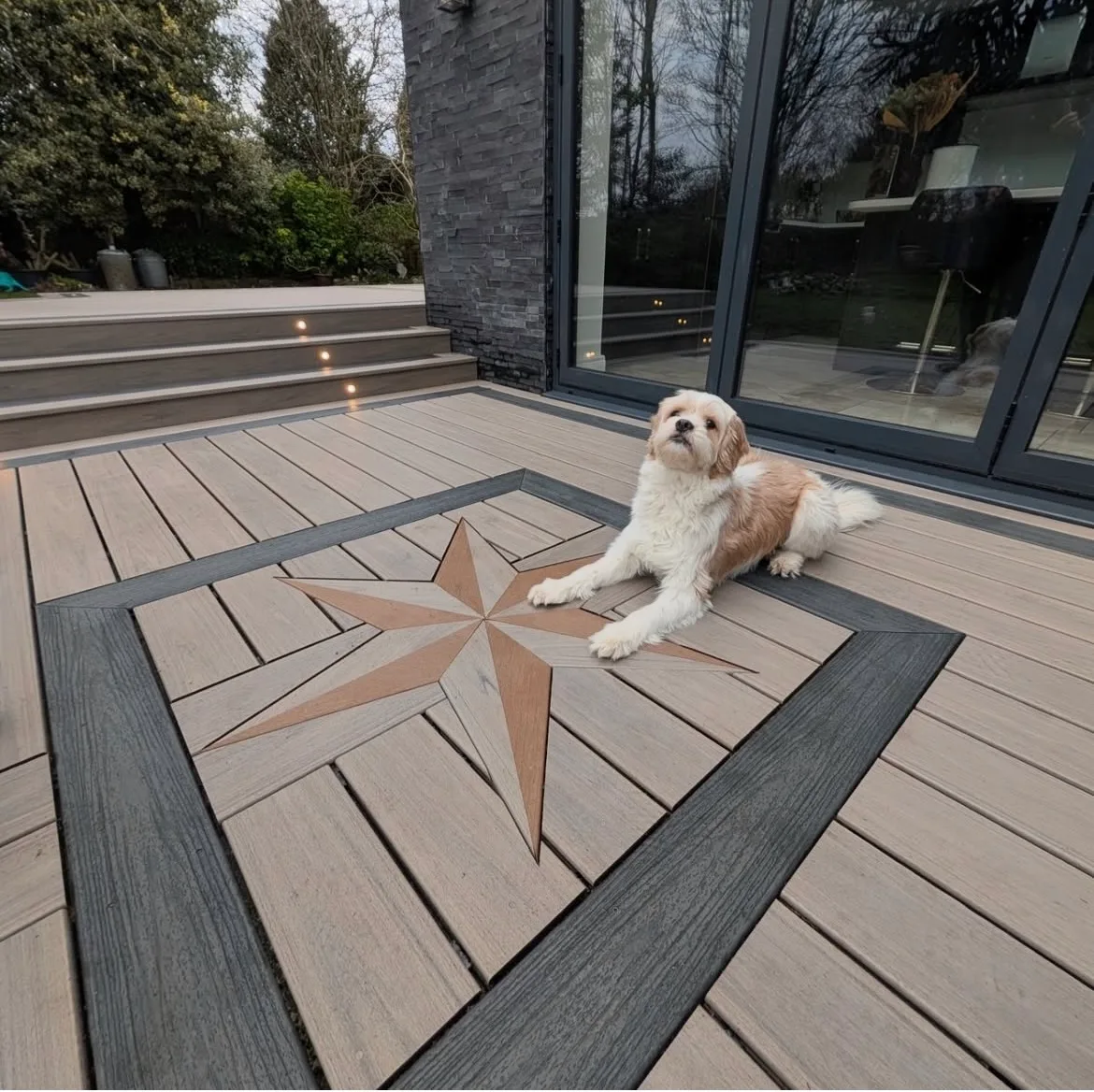 Composite deck with a compass star inlay in the boards and a dog resting on it beside a dark stone home