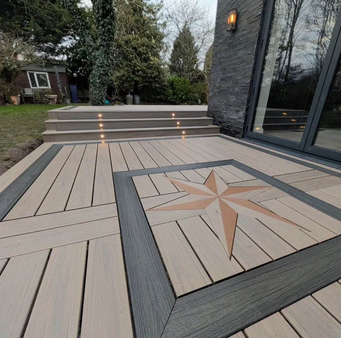 Composite deck with a compass star inlay and lit steps leading up to a modern dark home