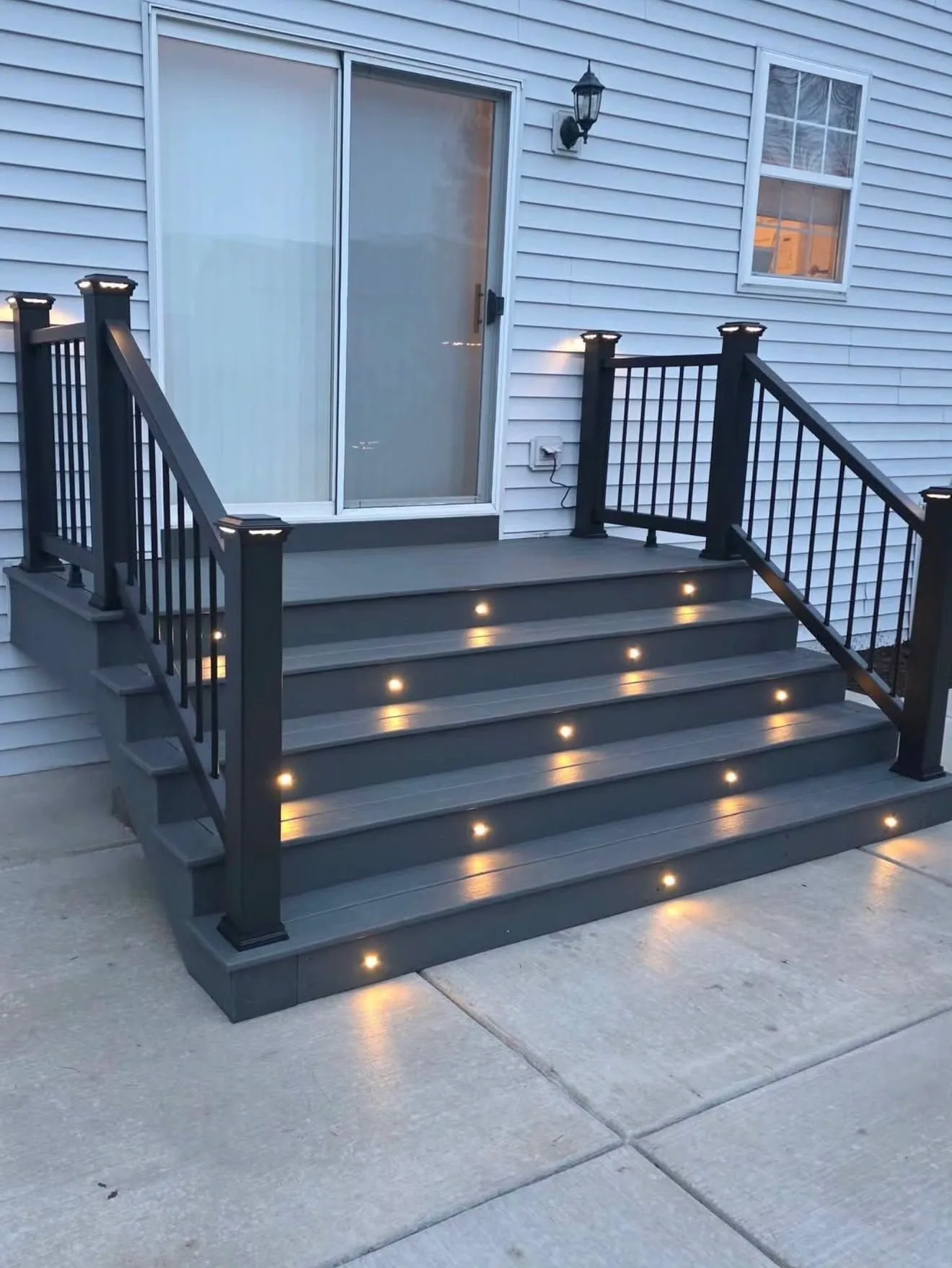 Dark composite steps with lights set into each riser and black railing at a blue sided home at dusk