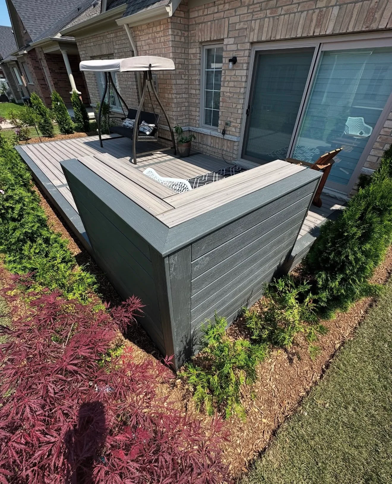 Grey composite deck with a built in bench and umbrella beside a brick home and red leaved shrubs